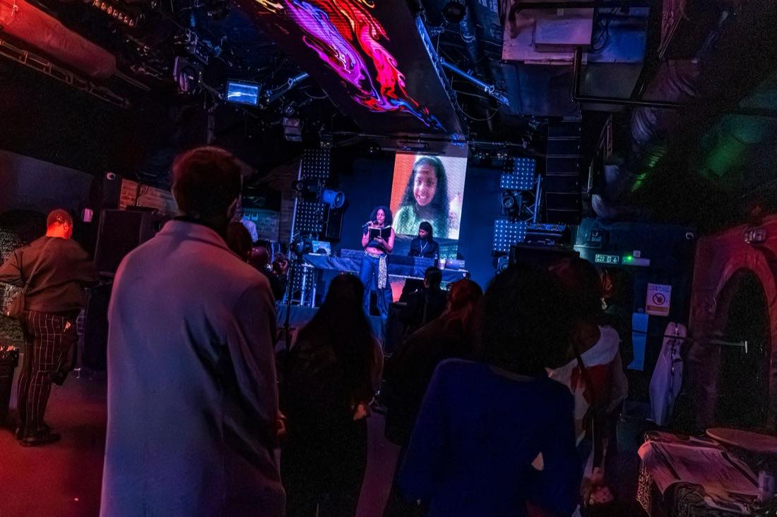 A poet reading from a book on stage, her face projected on the screen behind